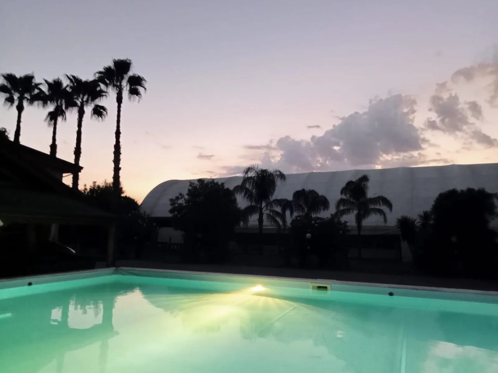 Piscina illuminata al tramonto, con le palme e la tensostruttura sullo sfondo